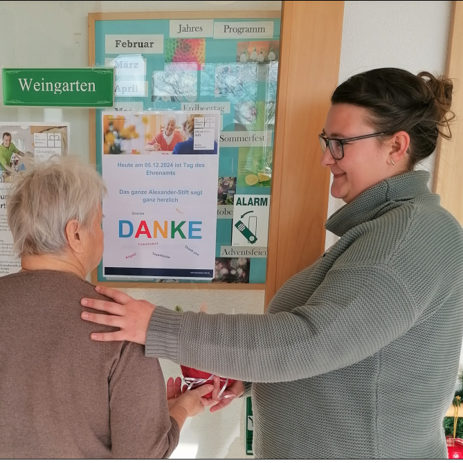  eine junge Frau mit brünetten, hochgesteckten Haaren klopft einer älteren, kurzhaarigen Dame auf die Schulter. Beide stehen vor dem Schwarzen Brett, an dem das Plakat hängt